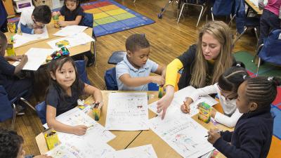 A teacher sits with her young students and points at a question on a sheet of paper.