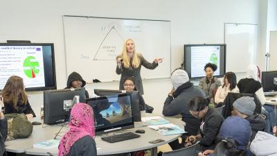 Instructor talks to crowded classroom in front of a board with a diagram drawn on it.