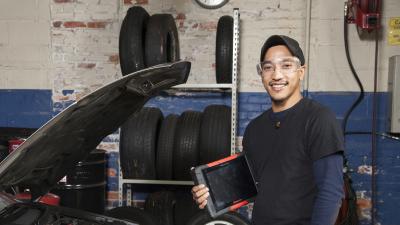 Automotive student in a garage holding flashlight and diagnostic tool in front of a car.