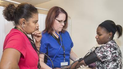A student checks the pulse of a patient while instructor looks on.