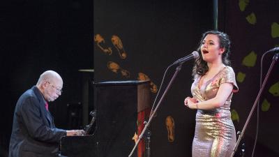 Singer performs at microphone with pianist in the background.