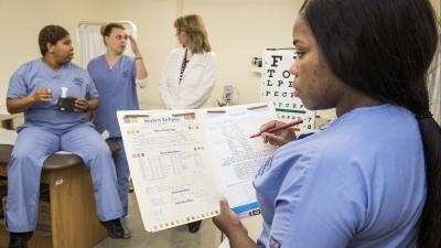 Clinical assistant fills out medical form with medical personnel in the background.