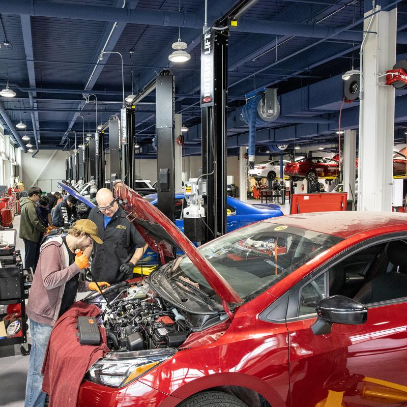 Student and instructor examine a car engine in a large garage.