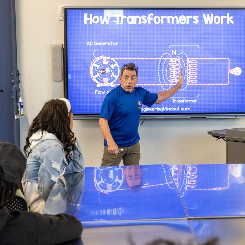 Instructor in classroom gestures toward a diagram on a digital screen.