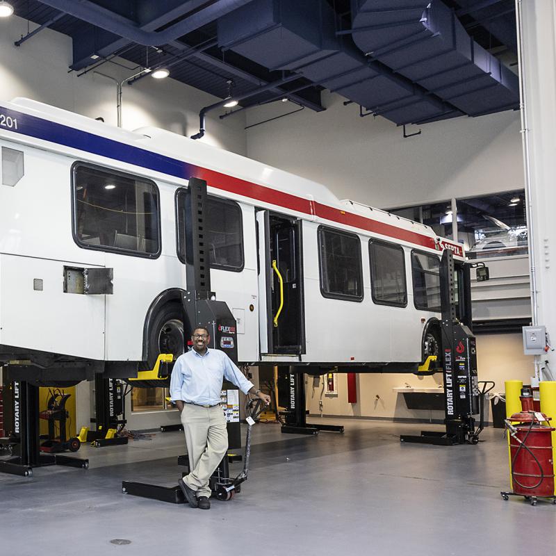 Instructor stands before a bus on a lift in a large garage.