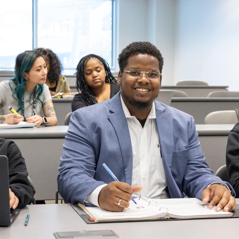 Student seated in a classroom witha notebook smiles for the camera.