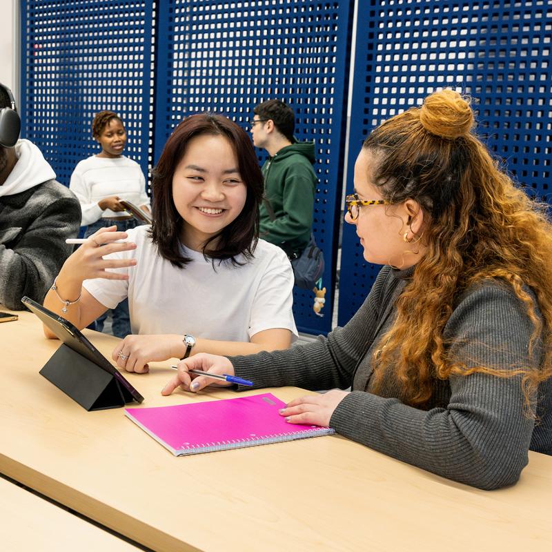 Two students in a campus common area discuss school work.