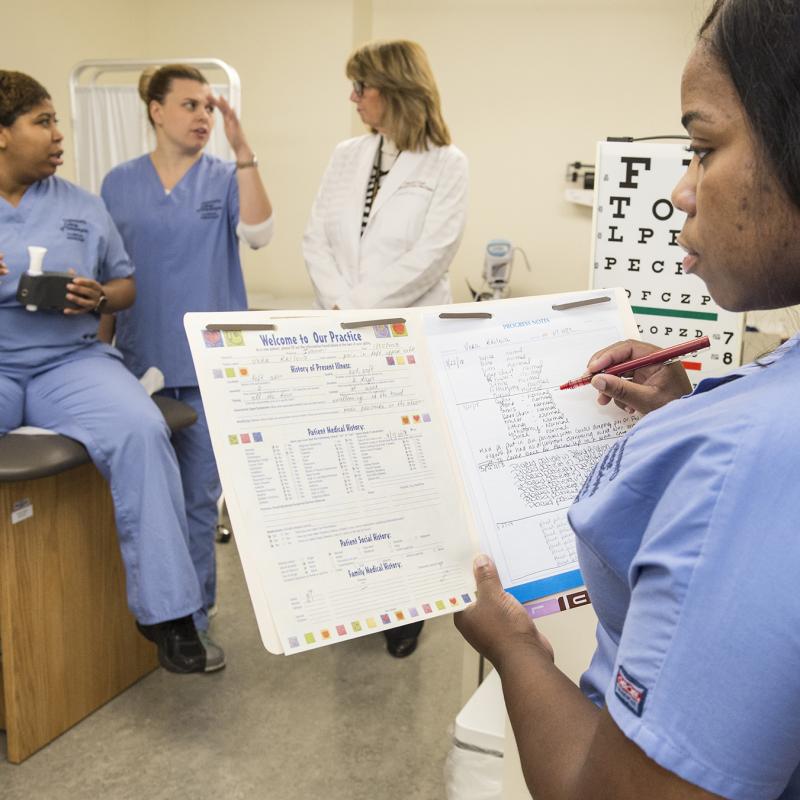 Assistant fills out medical records with medical personnel in the background.
