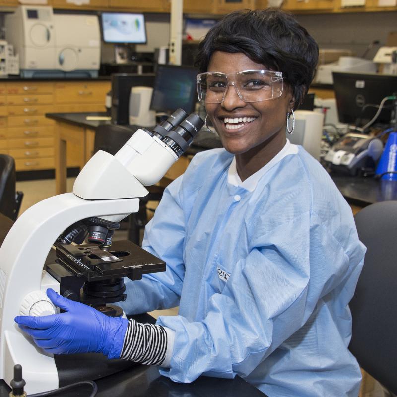 Lab tech at a microscope smiles for the camera.