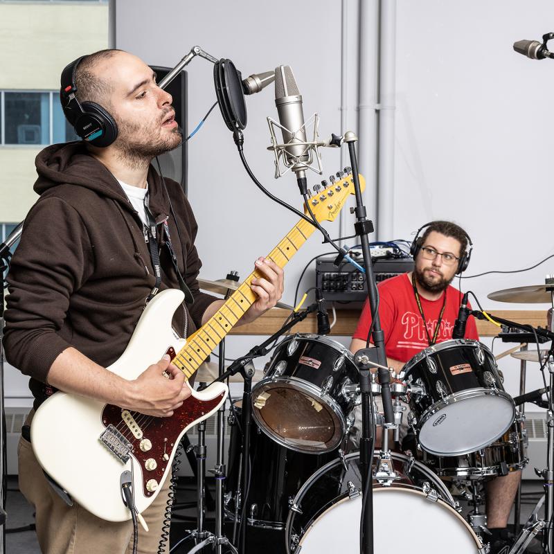 Guitarist sings as drummer watches in music studio.
