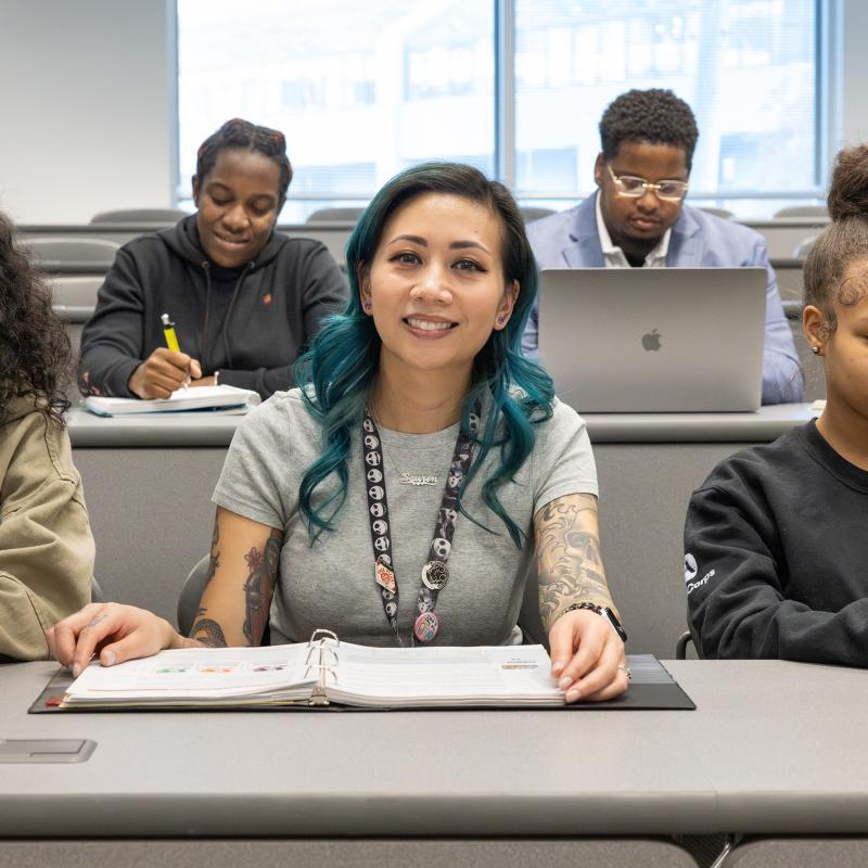 Student seated in classroom with a notebook, smiles for the camera.
