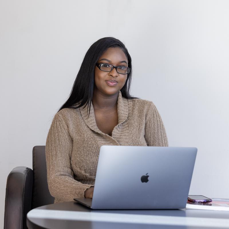 Woman seated at a table working on a laptop smiles for the camera.