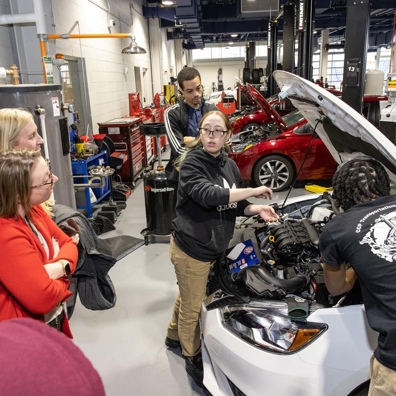 Female student showing people something under the hood of a car. 