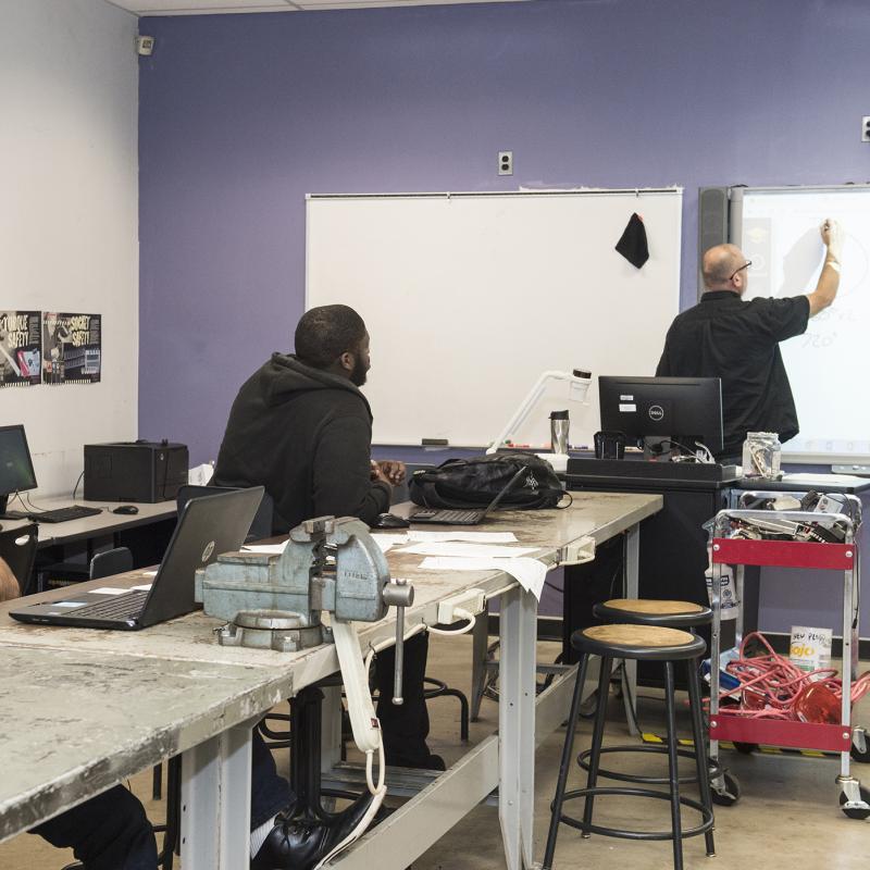 a man writing on a white board in front of a class of other people