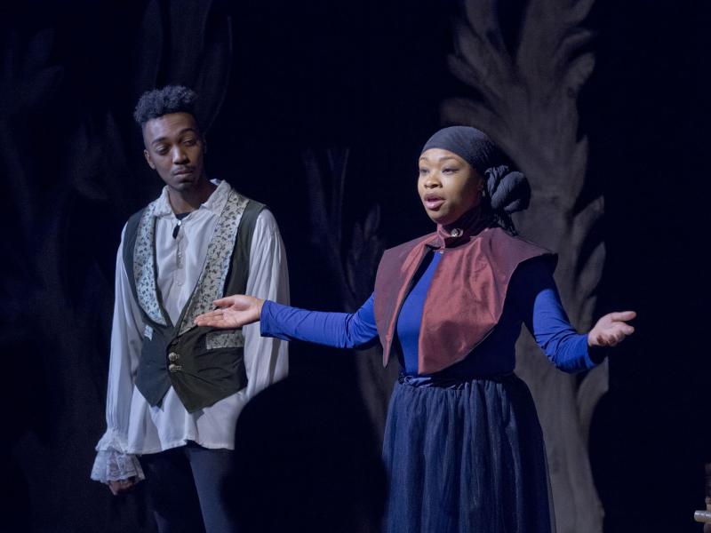 Actor speaks with outstretched arms while another actor looks on.