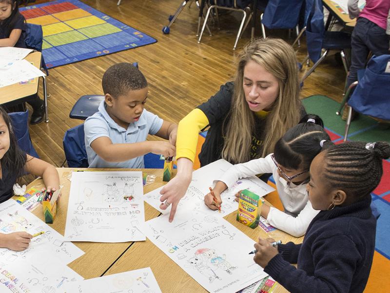 A teacher sits with her young students and points at a question on a sheet of paper.