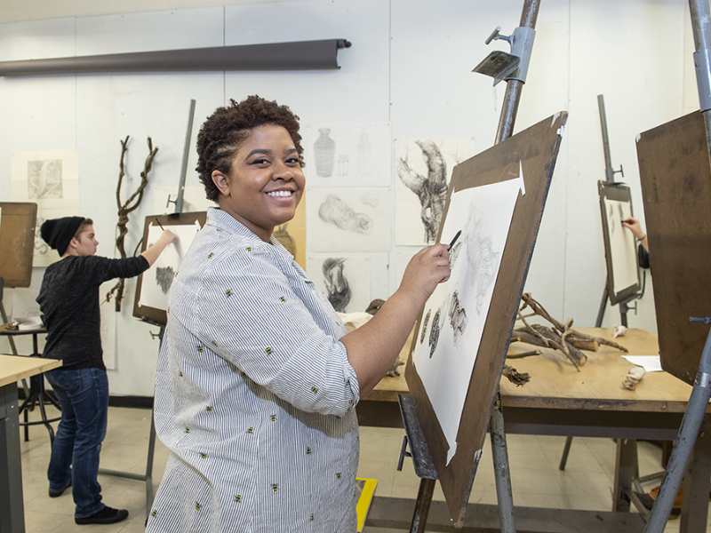 smiling student in art studio classroom drawing with charcoal at an easel