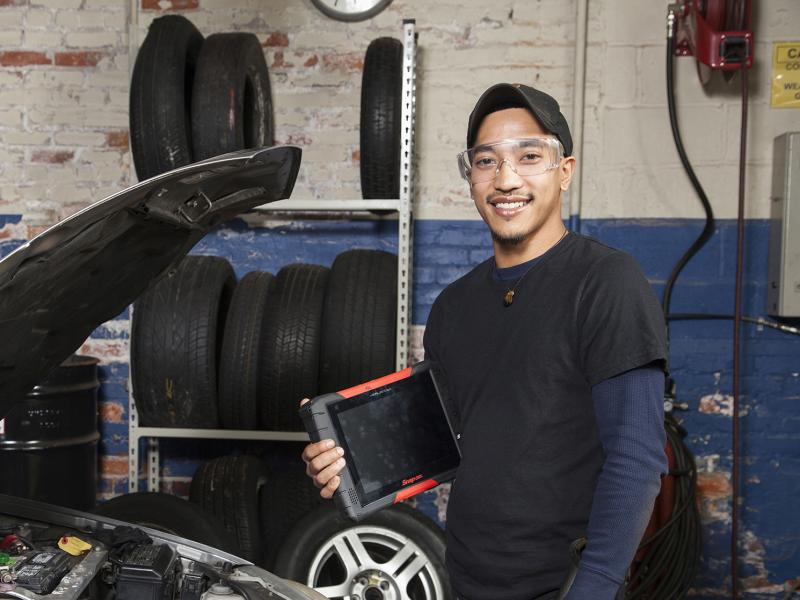 Automotive student in a garage holding flashlight and diagnostic tool in front of a car.