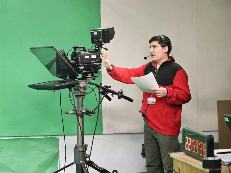 Student with broadcast camera and script speaks to people out of the frame.