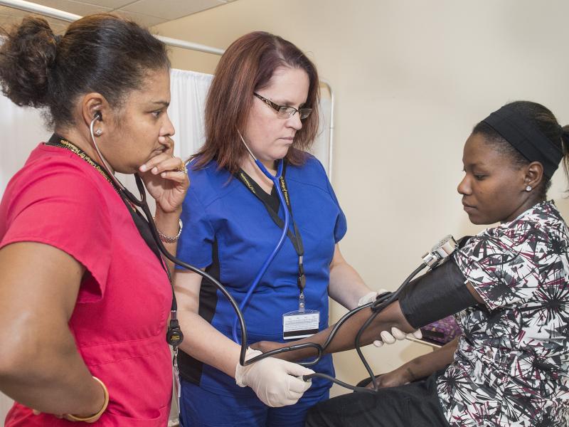 A student checks the pulse of a patient while instructor looks on.