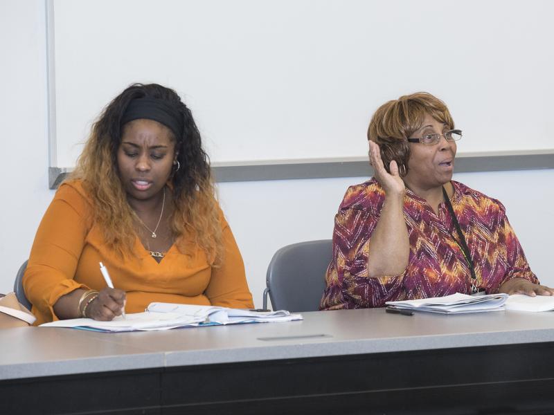 Student in classroom raises hand. Another student takes notes.