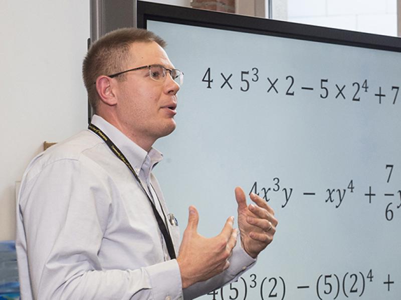 Instructor speaking next to a large digital screen with mathematic equations.