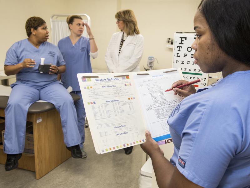 Clinical assistant fills out medical form with medical personnel in the background.
