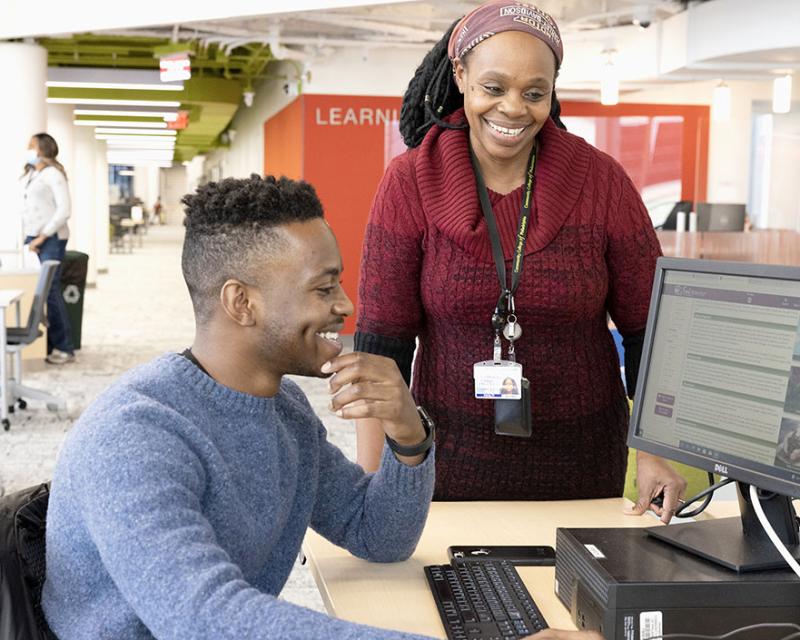 A student works at a Learning Lab computer while a faculty advisor smiles and guides them